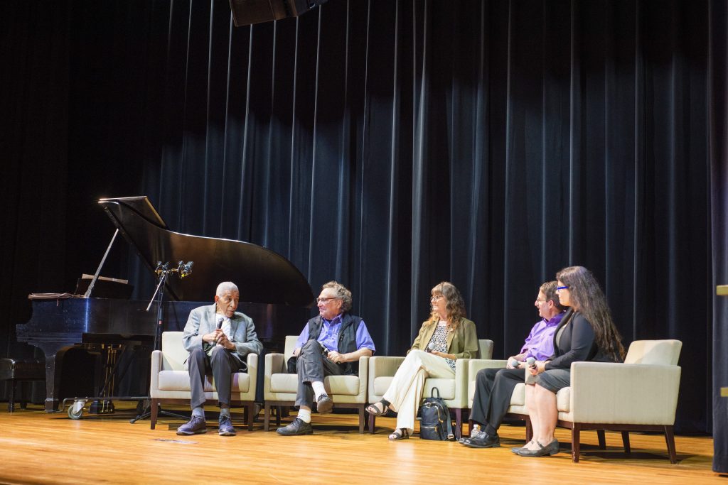 Panelists on stage listen to Norman speaking on stage